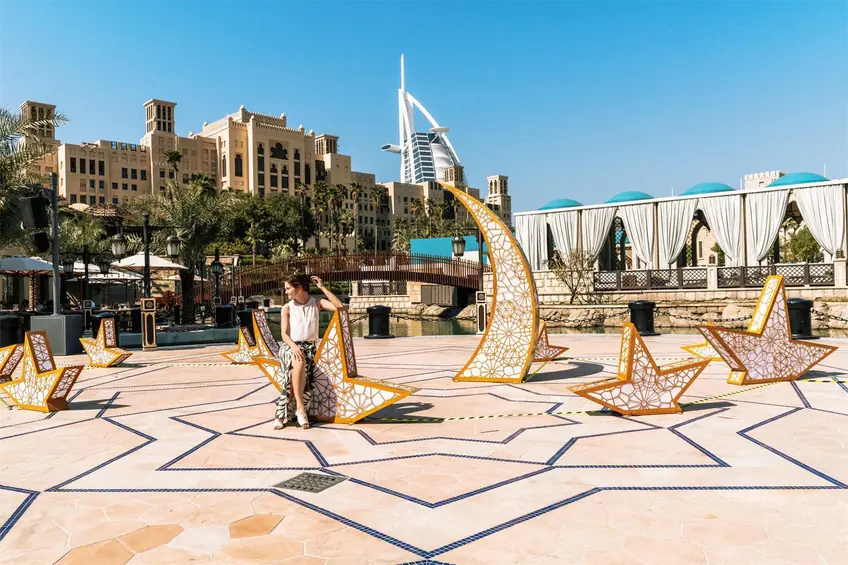 A woman relaxes by an artistic installation with the iconic Burj Al Arab in the background, set in Dubai's luxurious outdoor setting.